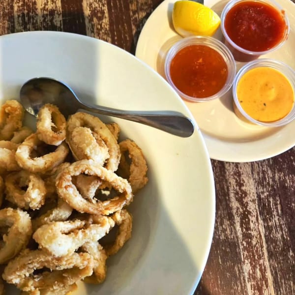 Crispy fried calamari in a white bowl with a spoon, served with three dipping sauces and a lemon wedge.