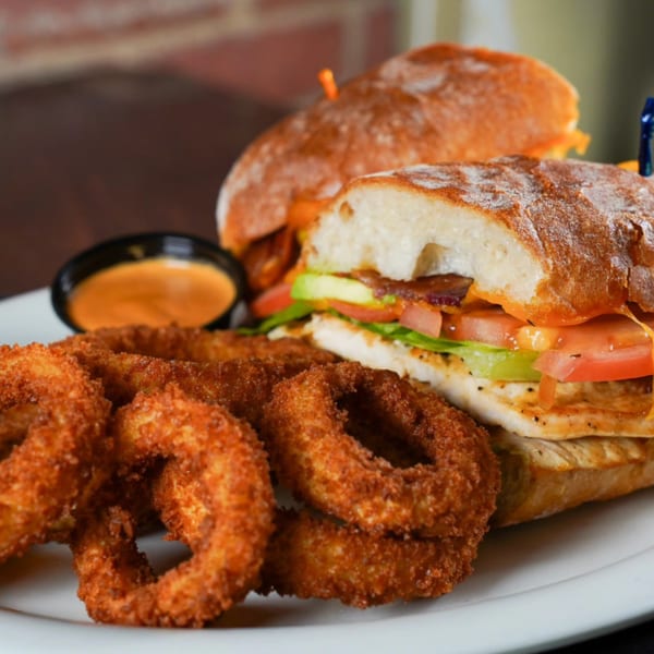Grilled chicken sandwich with bacon, lettuce, tomato, avocado, and a side of onion rings and dipping sauce.